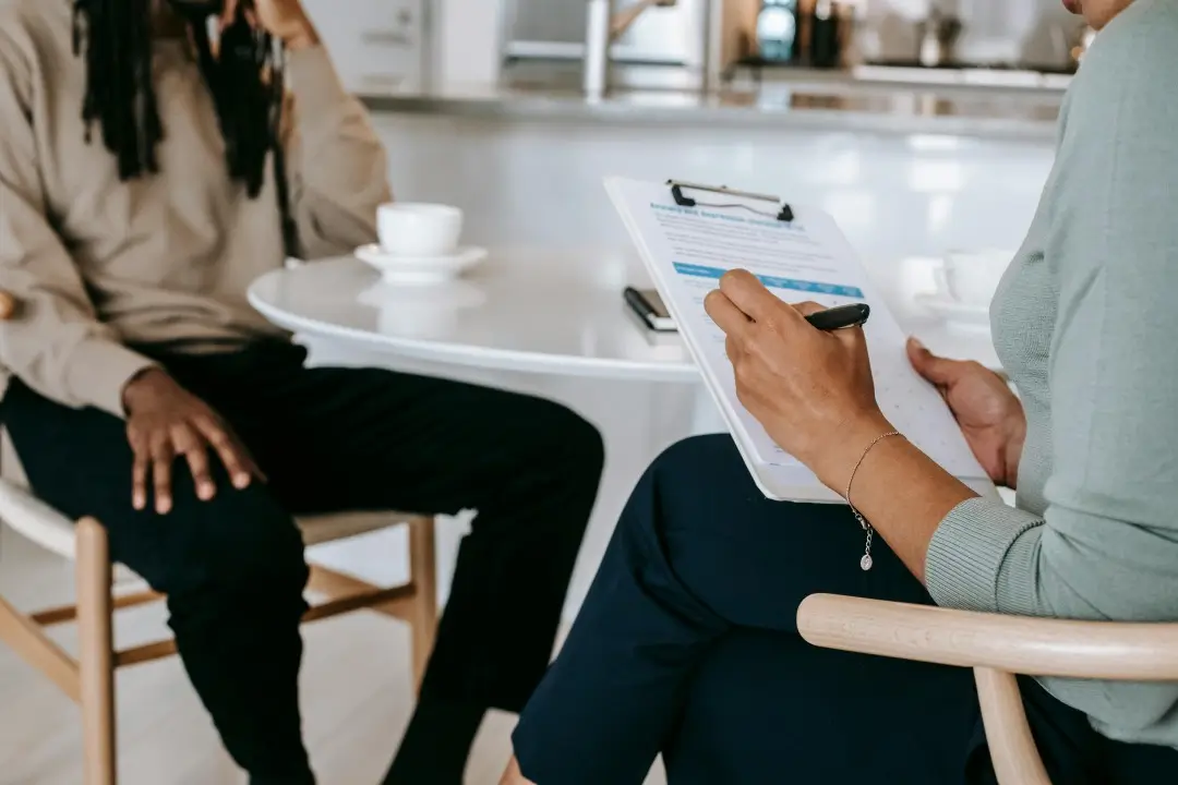 Close-up of a professional consultation where a woman is writing on a clipboard while seated across from a man in casual attire, both sitting at a round white table with coffee cups in a bright, modern office or kitchen setting.
