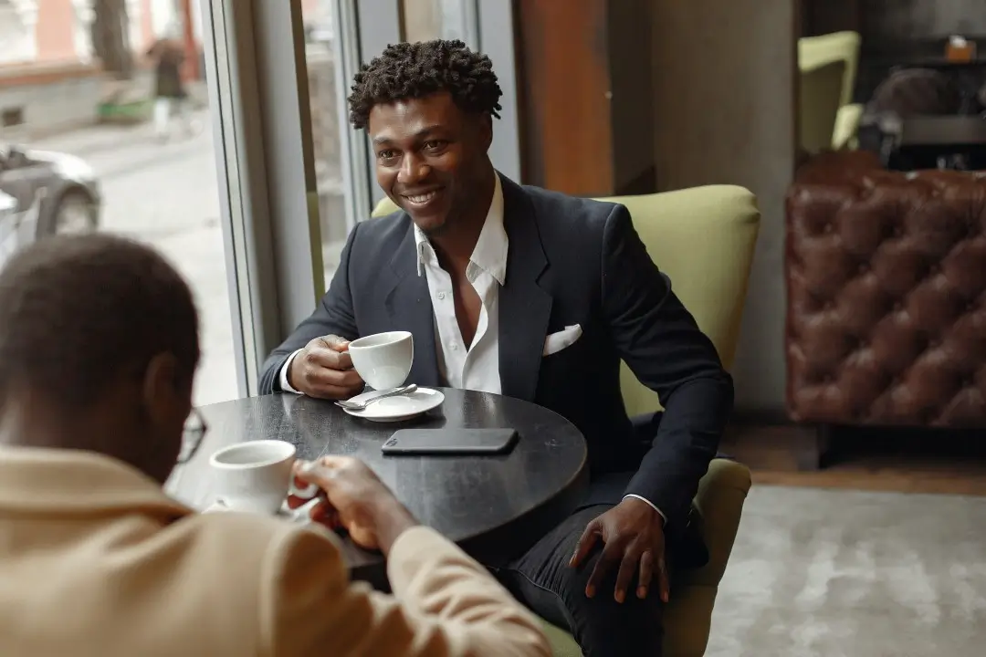 Two well-dressed men having a coffee meeting at a modern café, with one man smiling confidently in a dark suit while seated by a window, suggesting a professional or friendly conversation.
