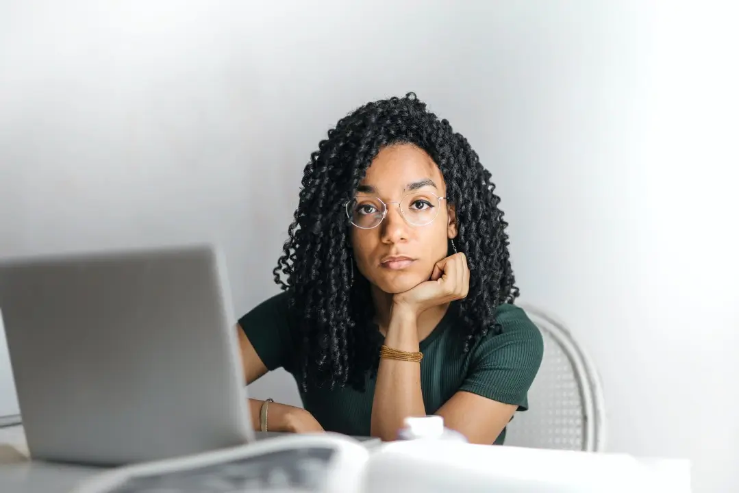 Young woman with curly hair and eyeglasses sitting at a desk with a laptop, resting her chin on her hand and looking directly at the camera with a neutral or contemplative expression in a minimal home office setup.